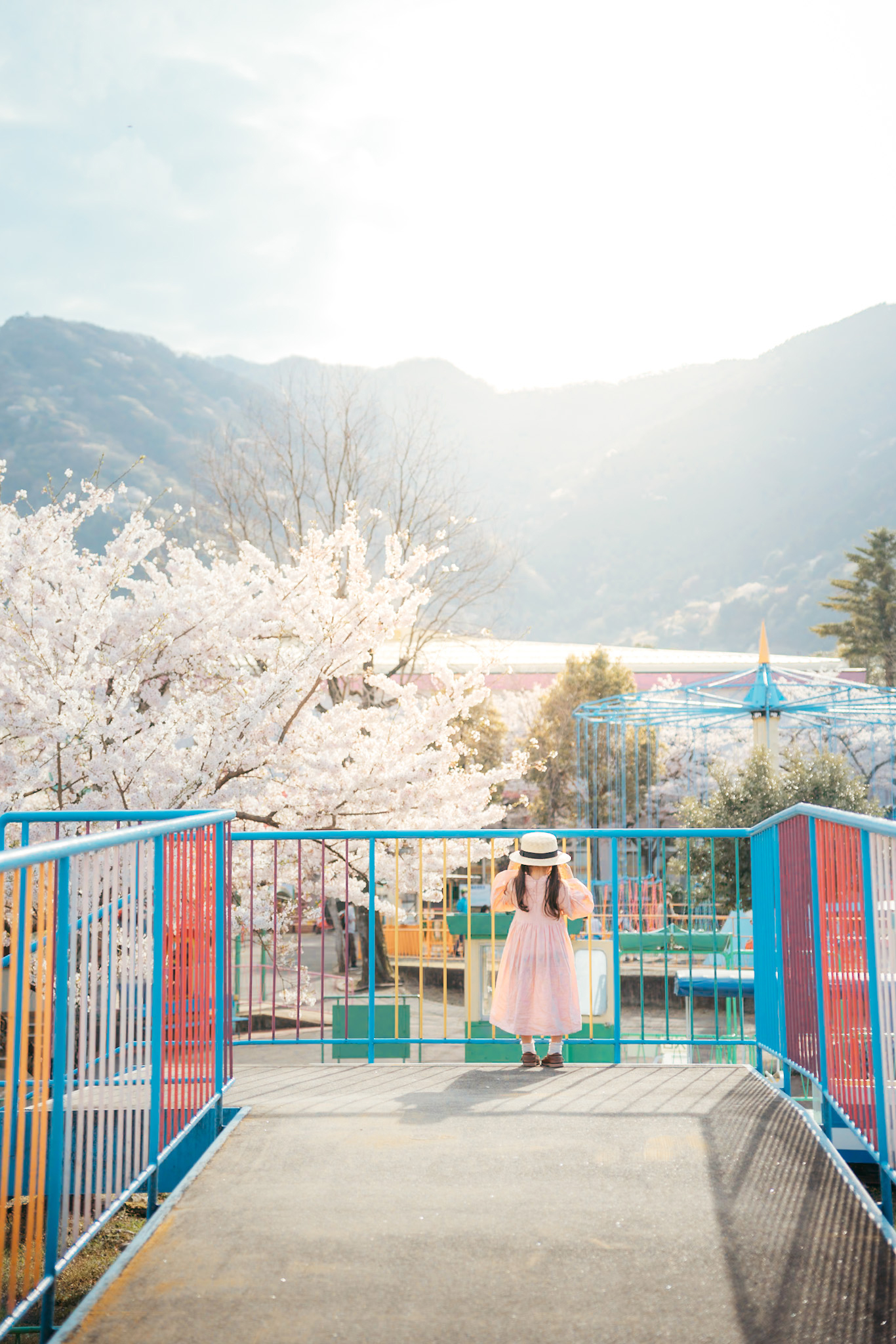 遊園地のカラフルな遊具と子供、その向こう広がる桜と山々が春の風景を彩る。遊び心と自然の美しさが融合した写真。