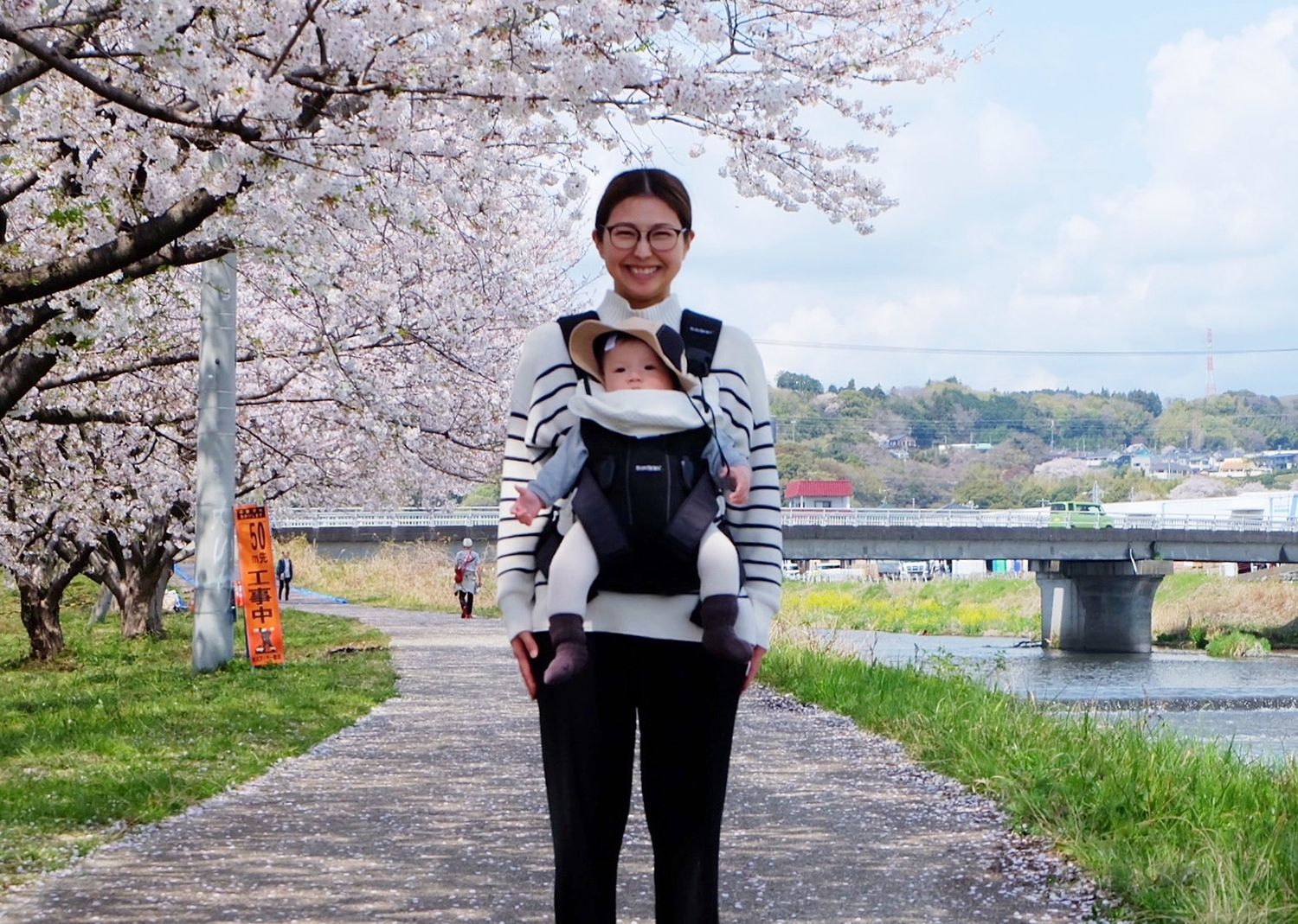 桜並木の道で赤ちゃんを抱えた人物が立つ春の風景。川沿いの橋や歩く人々が背景に映り、家族の時間と季節の彩りが重なる写真。